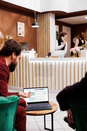 Les employés se réunissent à la réception de l'hôtel pour travailler sur la collaboration, formant des liens internationaux pour naviguer dans le paysage commercial mondial. Les entrepreneurs lisent des idées partagées sur ordinateur portable.