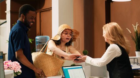 Photo for Hotel guests paying for room reservation, using credit card for payment on pos terminal at front desk reception. Couple on summer holiday making transaction upon their arrival. Handheld shot. - Royalty Free Image