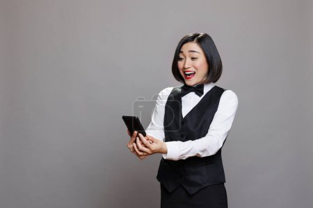 Photo for Excited happy asian waitress reading catering service positive reviews while holding smartphone. Young woman hotel receptionist in uniform checking good news message on mobile phone - Royalty Free Image