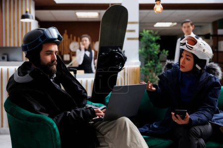 Photo for In hotel lobby young guests on couch plan skiing and snowboarding activities with digital devices. Couple using technology for online check-in at ski resort. Snowboard helmets and laptop available. - Royalty Free Image