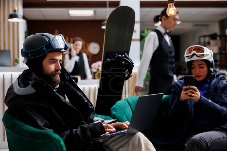 Photo for Two visitors sit comfortably using digital devices while snowboard equipment suggests nearby winter sports. Image showing man and woman surfing on laptop and mobile phone in ski hotel lobby. - Royalty Free Image
