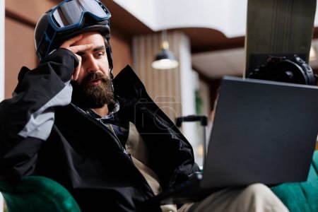 Caucasian man in winter clothes using laptop for research on snow activities at ski mountain resort. Male traveler with snowboard gear utilizing modern technology for online booking in hotel lobby.