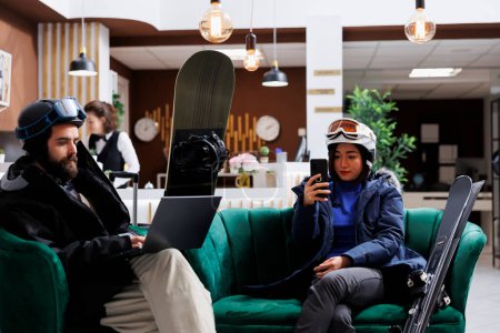 Photo for Young couple enjoys winter vacation by lounging in the ski resort lounge area with their digital gadgets. A man with a laptop and a lady with smartphone take a break from skiing and snowboarding. - Royalty Free Image