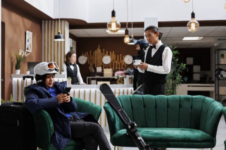 Photo for In hotel reception asian female tourist in winter attire uses digital device to explore snow gear and winter activities. Young waiter holding cup of coffee to woman with smartphone in lounge area. - Royalty Free Image