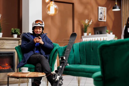 Happy female traveler wearing snow clothing seated in lounge area browsing on mobile phone for winter holiday activities. Asian woman with skiing equipment surfing the net in ski mountain resort.