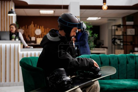 Photo for Young male traveler in winter gear sits in lounge area fine-tuning snowboard gear ready to explore the ski resort and try snowboarding. Man wearing winter jacket checks his wintersports equipment. - Royalty Free Image