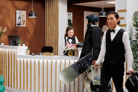 Female receptionist at front desk assists man with snowboarding equipment with accommodation reservation at winter resort. Professional bellboy taking luggage of male guest towards booked hotel room.