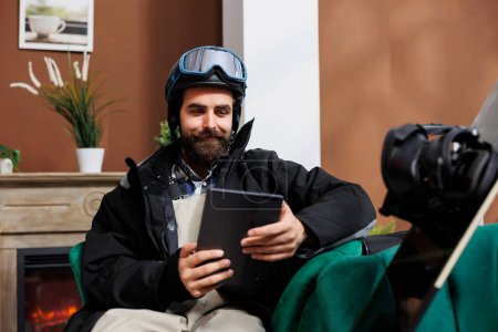 Foto de Viajero en equipo de invierno con dispositivo digital en la sala de estar de la estación de esquí, navegación y reserva de vacaciones de invierno. Hombre con barba sentado en un acogedor sofá, rodeado de equipo de snowboard. - Imagen libre de derechos