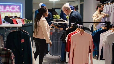 Foto de Diverse clients shopping in retail store, looking for black friday discounts. Customers browsing through clothing items on hangers and racks in fashion outlet, exploring seasonal sales. - Imagen libre de derechos