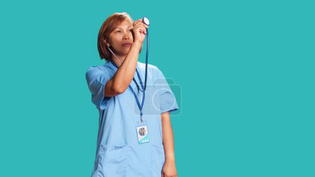Photo for Experienced asian healthcare specialist using stethoscope, measuring vital signs. Nurse isolated over studio background utilizing professional medical device to provide diagnostic - Royalty Free Image
