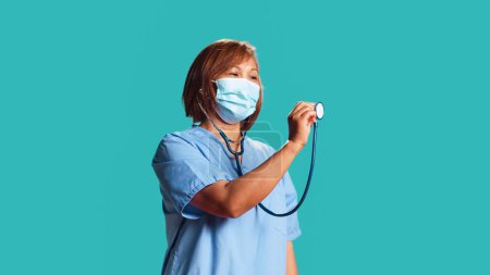 Photo for Asian experienced nurse doing mock up showing of properly using professional stethoscope. Knowledgeable clinic worker demonstrating medical gear usage, isolated over blue studio background - Royalty Free Image