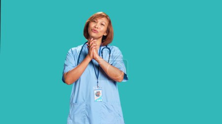 Pleading asian healthcare professional imploring hospital management for wage raise. Overworked nurse with hands folded in prayer gesture asking for days off, isolated over blue studio background