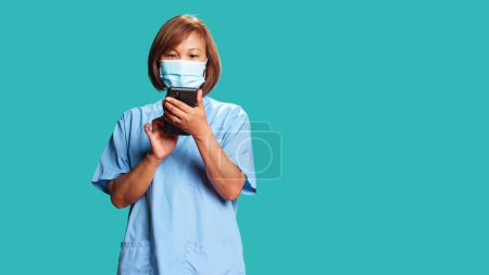 Happy relaxed nurse taking break between hospital shifts, texting friends, having fun online. Smiling woman wearing protective face mask using gadget isolated over blue studio background