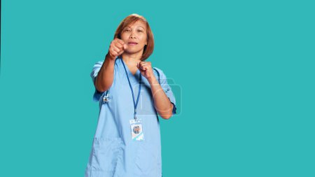 Photo for Happy playful asian nurse having fun pretending to fight, throwing punches. Funny high spirited healthcare specialist mock boxing while taking break from work, isolated over blue studio background - Royalty Free Image