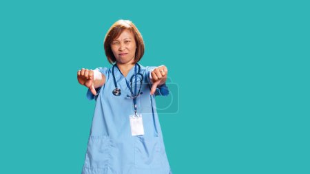Upset asian nurse showing thumbs down signs while at work, isolated over blue studio background. Dissapointed BIPOC healthcare professional doing frenetic disapproval gestures