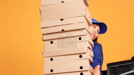 Téléchargez les photos : Coursier surmené submergé par une grosse pile de boîtes à pizza, attendant que le client réponde à la porte. Cycliste fatigué isolé sur fond de studio livrant ordre alimentaire lourd, gros plan - en image libre de droit