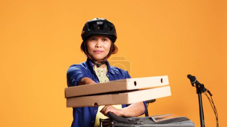 Foto de Primer plano de la mensajera aislada sobre fondo de estudio naranja trayendo pizza al cliente. ciclista asiático entrega comida para llevar al cliente, sacando cajas de comida de la bolsa térmica - Imagen libre de derechos