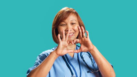 Téléchargez les photos : Gros plan de joyeux infirmier qualifié BIPOC faisant forme de symbole cardiaque avec les mains. Employé de la clinique montrant un geste d'amour au travail, isolé sur fond de studio bleu - en image libre de droit
