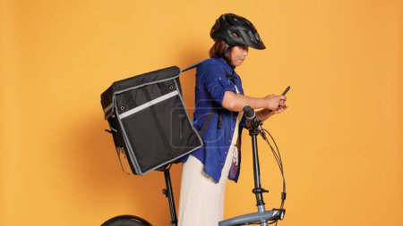 Téléchargez les photos : Cycliste asiatique livrant des plats à emporter aux clients, imputant les données de localisation suivantes dans l'application téléphonique. Courrier femme vélo, port casque de sécurité et sac à dos thermique, isolé sur fond de studio - en image libre de droit