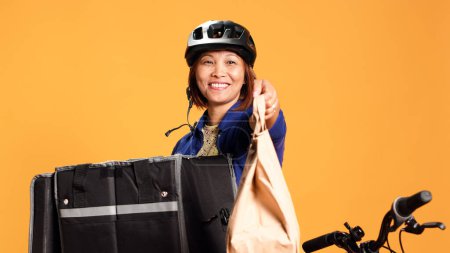 Photo for Handheld close up shot of BIPOC woman taking takeaway meal bag out of professional thermal backpack. Friendly courier isolated over orange studio background handing client food delivery order - Royalty Free Image