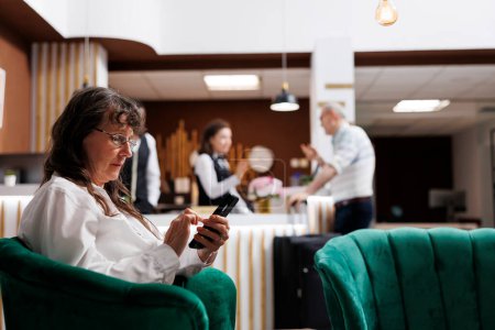 Caucasian senior female traveller relaxing on comfy sofa with smartphone in hotel lounge area. Retired senior woman looking for enjoyable holiday activities on her mobile device in beautiful resort.