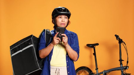 Confused asian woman looking around for directions, unsure of client location whereabouts. Professional courier wearing thermic backpack riding bike to deliver food, isolated over studio background