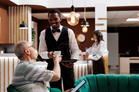 Afrikanisch-amerikanischer Mitarbeiter serviert einem älteren männlichen Touristen an der Rezeption eines Luxushotels Kaffee mit Tablette. Älterer Mann hält ein digitales Gerät in der Hand und erhält eine Tasse Tee vom Hotelkellner im Aufenthaltsraum.