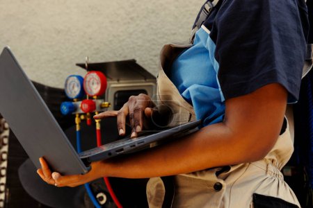 Photo for Specialist african american mechanic using laptop to look online for new capacitor after finding condenser malfuntioning internal component during business owner comissioned maintenance - Royalty Free Image