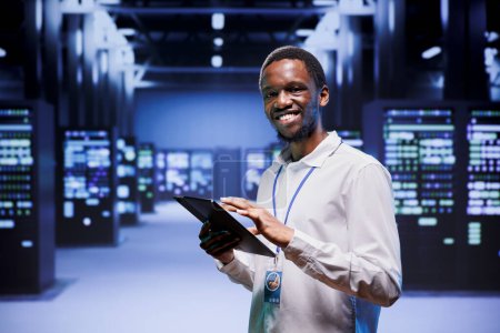 Porträt eines afrikanisch-amerikanischen Administrators vor Serverclustern im Hochleistungsrechenzentrum. Techniker überwacht systemübergreifend den Energieverbrauch, um einen optimalen Betrieb zu gewährleisten