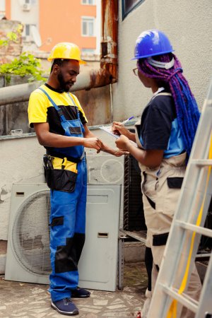 Photo for Efficient technicians repairing and maintaining condenser heating and ventilation. Knowleadgeable engineers hired for outdoor air conditioner routine checkup, writing report on clipboard - Royalty Free Image