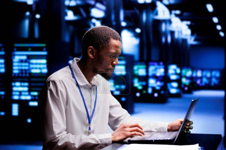 African american IT developer in data center using laptop to set up configuration management tools that enable automatic failover and load balancing, preventing server cabinets system crash