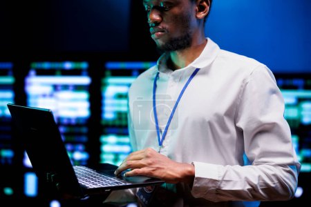 African american IT developer using laptop to examine server mainframes providing vast computing resources. Data center enabling AI to process massive datasets for complex machine learning operations