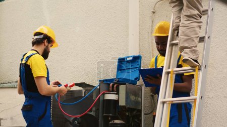 Spezialisten messen Freon-Pegel in HLK-Anlage mit Druckindikatoren, während afrikanisch-amerikanischer Mitarbeiter von Klappleiter absteigt, nachdem er Dachkondensator überprüft hat