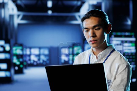 Skilled programmer in data center uses laptop to prevent system overload during peak traffic periods. Experienced professional in server room ensuring enough network bandwidth for smooth operations