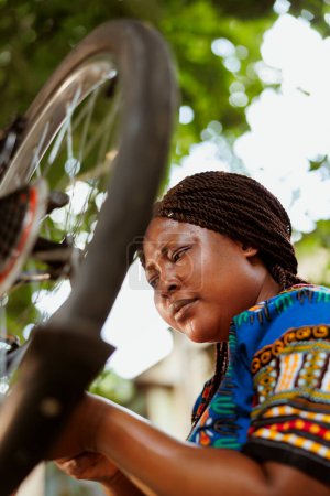 Foto de Mujer afroamericana activa trabajando en rueda de goma de bicicleta rota con equipo profesional en el patio de casa. Comprometida deportista negro mujer servicio de bicicleta fuera para el verano de ocio en bicicleta. - Imagen libre de derechos