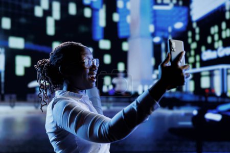 Téléchargez les photos : Femme afro-américaine utilisant un téléphone portable pour prendre des selfies tout en se promenant dans la ville la nuit. Citoyen utilisant un smartphone pour prendre des photos d'elle-même tout en se promenant à l'extérieur dans des rues vides - en image libre de droit
