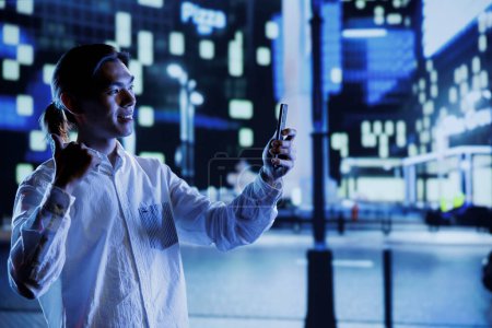 Photo for Man outside in city at night, using mobile phone to take selfie. Citizen using smartphone to take pictures of himself showing ok hand gesture sign while on empty streets illuminated by lamps - Royalty Free Image
