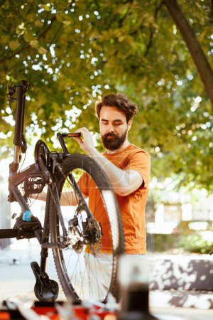 Photo for Healthy sports-loving man securing bike wheel for outdoor leisure cycling. Active young caucasian male cyclist doing yearly maintenance and adjusting bicycle tire in yard during the summer. - Royalty Free Image