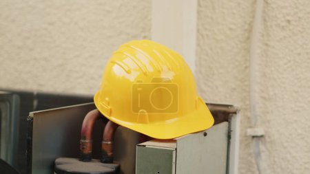 Extreme close up of yellow safety hardhat on top of out of order external air conditioner unit in need of repair. Protective professional engineering gear left on opened HVAC system