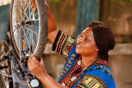 Téléchargez les photos : Jeune femme active effectuant l'entretien annuel et ajuster les pneus de vélo dans la cour pendant l'été. Femme africaine américaine sportive saine sécurisant roue de vélo pour le cyclisme de loisirs de plein air. - en image libre de droit