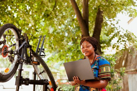 Foto de Mujer afroamericana dedicada sana que busca en el ordenador portátil para arreglar la bicicleta moderna rota. La ciclista femenina activa garantiza que los componentes de la bicicleta sean seguros para el ciclismo de ocio al aire libre de verano. - Imagen libre de derechos