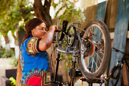 Active african american lady adjusts bike pedal and fine-tunes the gears as yearly summer maintenance. Sports-loving black woman servicing and fixing modern bicycle in home yard.