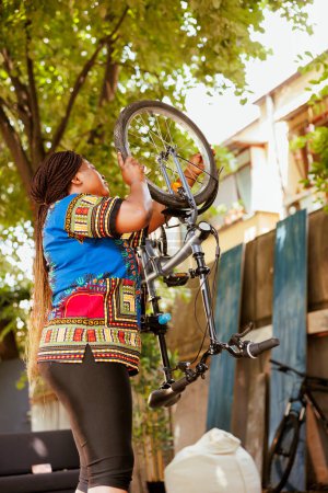 Die aktive, sportbegeisterte Afroamerikanerin sorgt dafür, dass das Rad für das sommerliche Freizeitradeln sicher ist. Gesunde schwarze Radfahrerin befestigt Fahrradreifen sicher im heimischen Hof.