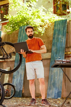 Téléchargez les photos : Homme caucasien dévoué sain utilise son ordinateur portable pour réparer cassé vélo moderne dans la cour de la maison. Image montrant un jeune cycliste portant un mini-ordinateur pour l'entretien du vélo à l'extérieur. - en image libre de droit