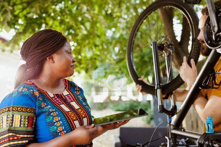 Photo for Active black woman enjoys browsing internet while active man fixes and maintains bicycle. Smiling african american female holding digital device as sport-loving caucasian male inspects bike tire - Royalty Free Image