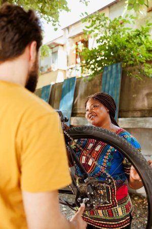 Photo for Sports-loving black woman checking and maintaining bicycle for yearly summer cycling. Active young caucasian man helps healthy energetic african american woman mend damaged bike wheel outside. - Royalty Free Image
