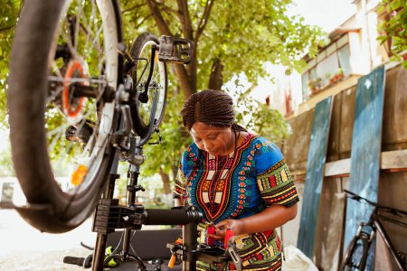 Foto de Energetic africana amerciana ciclista inspeccionar cuidadosamente equipo especializado para el mantenimiento de la bicicleta en el exterior. Mujer negra amante de los deportes en busca de herramienta de trabajo en el patio de casa dedicada al verano - Imagen libre de derechos