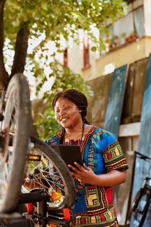 Téléchargez les photos : À l'extérieur active femme noire vibrante explorer l'Internet pour l'entretien du vélo. Femme cycliste afro-américain inspecter et réparer soigneusement vélo avec tablette numérique. - en image libre de droit