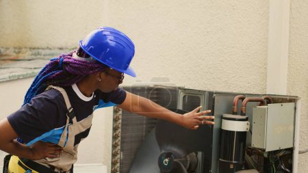 Trabajador afroamericano contratado para reparar el aire acondicionado roto, probando ventilador defectuoso ralentizado por la suciedad. Wireman investiga sistema de hvac abierto para comprobar si hay cableado incorrecto