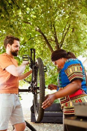 Photo for Sporty multiethnic couple inspecting and repairing bike tire and chain in home yard. Healthy and active two persons working outdoors on bicycle maintenance with expert work tool. - Royalty Free Image
